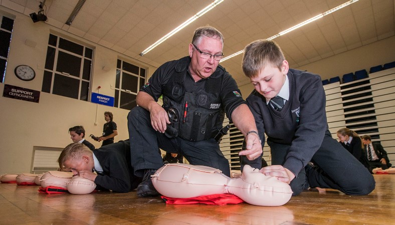 Ysgol Emrys ap Iwan, Abergele. Year 7 & 8 students learning First Aid and how to do CPR in a Restart Heart Campaign. Core Chapman, 11 gets some hints from PC John Wheway